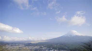 Fuji Dağı, parlak mavi gökyüzünün altında karla kaplı zirvesini sergiliyor. Sakin manzara yakındaki kasabaları ve yemyeşil alanları gözler önüne seriyor..