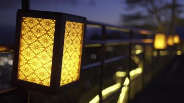 Sıcak turuncu Japon fenerleri alacakaranlıkta yürüyüş yolu boyunca hafif bir parıltı yayarak şehrin arka planında parlayan gökyüzü gibi sakin bir atmosfer yaratıyor..