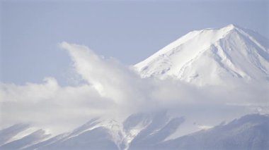 Sersemletici bir Fuji dağı manzarası açık mavi gökyüzünün altında görkemli bir karla kaplı zirve sergiliyor. Yumuşak bulutlar tembelce sürüklenir