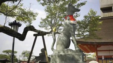 Zarif bir tilki heykeli gururla duruyor Fushimi Inari geleneksel Japon tapınağında, canlı kırmızı bir eşarpla süslenmiş..