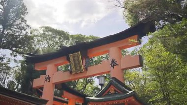 Canlı bir kırmızı torii kapısı ağaçların arasında görkemli bir şekilde duruyor, kutsal bir bölgenin girişini sembolize ediyor. Metin çevirisi: 