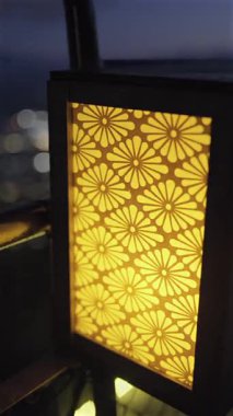 Güzel bir çiçek feneri balkona sıcak bir ışık yayar ve şehir ışıkları akşam saatleri boyunca parıldarken rahat bir atmosfer yaratır. Dikey Video