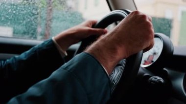 Bir adamın ellerini direksiyona koyup yağmurda yolda araba sürmesi.