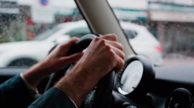 Bir adamın ellerini direksiyona koyup yağmurda yolda araba sürmesi.