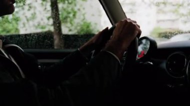 Bir adamın ellerini direksiyona koyup yağmurda yolda araba sürmesi.
