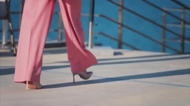 Pembe elbiseli, yüksek topuklu bir kadın sakin bir su manzarasıyla manzaralı bir terasta yürüyor.