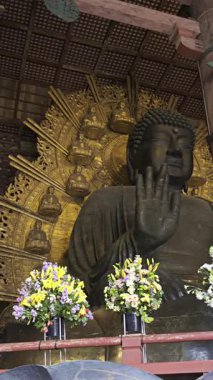 Nara, Japonya - 10 Nisan 2025: Todai-ji Budist tapınağı içinde büyük bir Buda heykeli ziyaretçileri karşılıyor. Heykelin altındaki sunağı renkli çiçekler süslüyor. Büyük Buda Salonunun içi