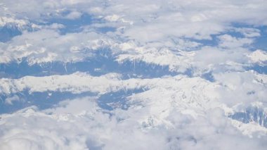 Karlı dağların üzerindeki beyaz, tüylü bulutların hava manzarası bir uçak penceresinden görülüyor.