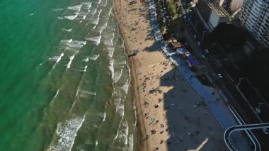 İspanya 'nın Alicante kentindeki plajda gün ışığında rahatlayan insanların bulunduğu kıyı şeridi boyunca giden arabaların insansız hava aracı görüntüsü.