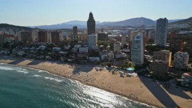 İspanya 'nın Benidorm plajında gün ışığında dinlenen insanların bulunduğu kıyı şeridi boyunca binaların insansız hava aracı görüntüsü.