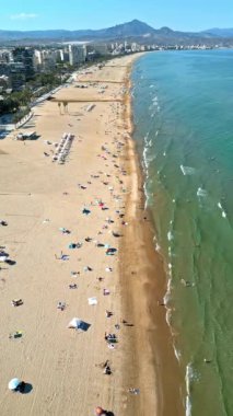 İspanya 'nın Alicante kentindeki plajda gün ışığında dinlenen insanların bulunduğu sahil boyundaki binaların insansız hava aracı görüntüsü. Dikey