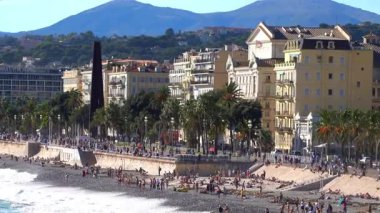 Güzel, Fransa - 3 Temmuz 2025: Palmiye ağaçları, tarihi binalar ve çakıl taşlı kıyı şeridinin keyfini çıkaran insanlarla dolu Gezinti Güvertesi des Anglais geniş görüş alanı