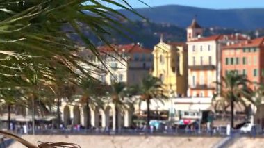 Nice 'in plajının panoramik manzarası önünde palmiye yaprakları, arka planda tepeleri olan pastel renkli binalar.