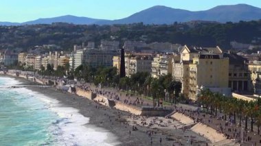 Turkuaz dalgalar, Gezinti Güvertesi des Anglais 'in çakıl taşı plajı boyunca çarpışıyor. Renkli binalar ve deniz kenarında palmiye ağaçları var.