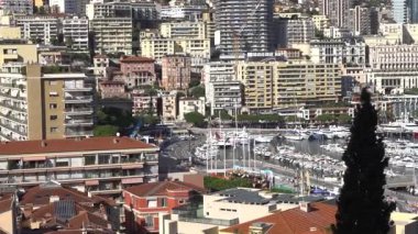 Monte Carlo, Monako - 4 Temmuz 2025: Çevredeki dağların çerçevelediği binalar, yüksek kuleler ve lüks yatlarla dolu hareketli marina manzarası
