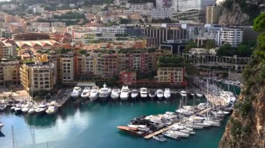 Monaco 'daki Port de Fontvieille' in yüksek açılı manzarası renkli binalar, lüks yatlar ve çevresindeki dağlık manzaraları gösteriyor.