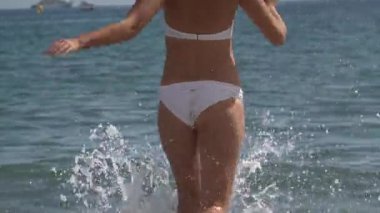 Beyaz bikinili bir kadın diz boyu sörf yapıyor. Parlak, güneşli ışıklar salıyor.