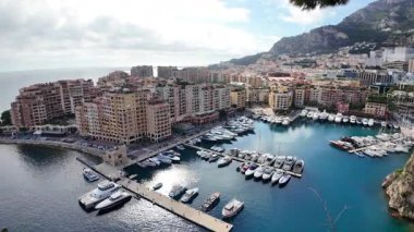 Monaco 'daki Port de Fontvieille' in yüksek açılı manzarası renkli binalar, lüks yatlar ve çevresindeki dağlık manzaraları gösteriyor.