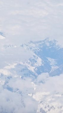 Karlı dağların üzerindeki beyaz, tüylü bulutların hava manzarası bir uçak penceresinden görülüyor. Dikey