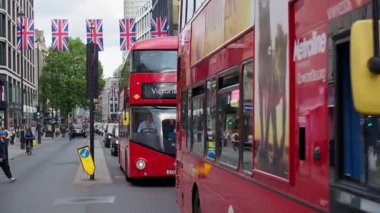 Londra, İngiltere - 12 Mayıs 2025: Klasik kırmızı çift katlı otobüsler, arabalar ve Union Jack bayrakları altında sokaklarda hareket eden insanlar