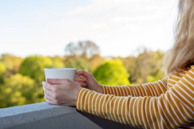 Yüksek binadaki balkonun kenarında arka planda bulanık ağaçlarla birlikte bir fincan sıcak içecek..