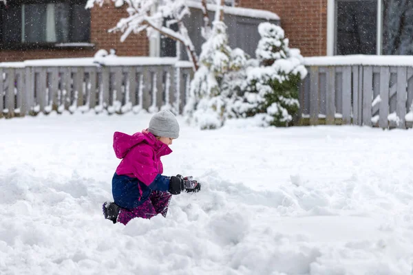 Little girl playing with snow outdoor near house in winter. Wintertime fun activities for children. Kid sitting in snow, doing snowballs