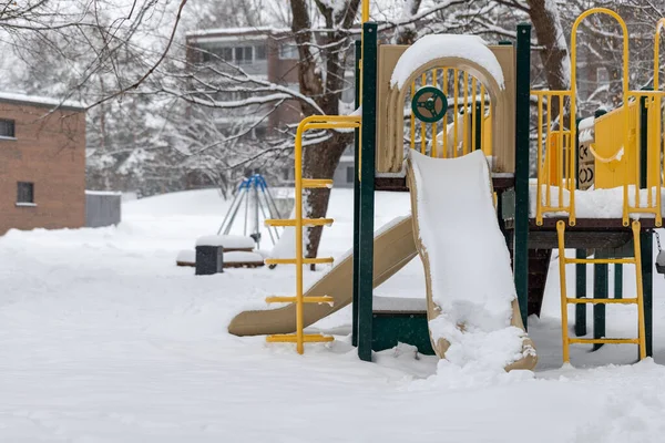 Playground with slide in park without children near houses, covered with snow in winter season