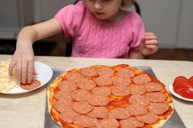 Küçük kız pepperonili pizza yapıyor. Evdeki mutfakta yemek pişiren küçük bir çocuk. Yemek hazırlama.