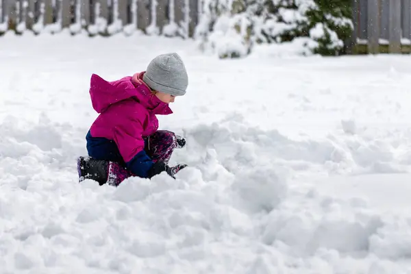 Little child playing with snow outdoor near house in winter. Wintertime fun activities for children. Kid sitting in snow, making snowballs