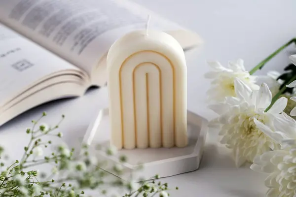 stock image White soy wax candle on a table with a book and flowers. Home decor. Cozy interior.