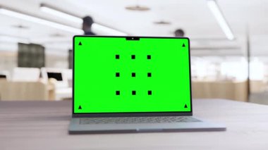 Modern bir ofiste ahşap bir masanın üzerinde yeşil renkli bir ekran ve arka planda bulanık bir kişi olan bir laptop..