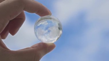 Bir el, mavi gökyüzünü ve bulutları yansıtan, görüşü, berraklığı ve küresel perspektifi sembolize eden berrak bir kristal küreyi tutar..
