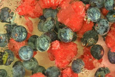 Blueberries and raspberries in liquid with bubbles. Colourful ripe bilberries and raspberries in water. Close-up of fresh berries in water background. Top view, flat design.