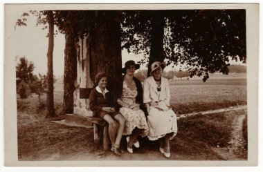 The CZECHOSLOVAK REPUBLIC - CIRCA 1930 'lar: Klasik fotoğraf, kadınların zarif elbiseler giydiğini ve Paris' te kadınların büyük şapka taktığını gösteriyor. Kadınlar ve çocuk dışarıda poz verip bankta oturuyorlar. Retro siyah beyaz fotoğraf. Çember 1930 'lar