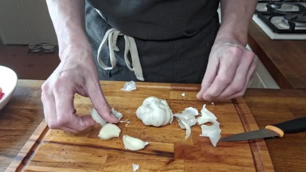 クックはニンニクのクローブを剥がします マンの手は木製の台所の切断板の上にニンニクのストリップを皮をむきます 家庭料理 グルメのコンセプト — ストック動画
