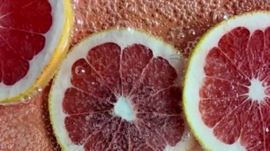 Suyun içinde kırmızı greyfurt dilimleri. Köpüklü suda kanlı kırmızı greyfurtlar. Meyve, vitamin, içki ve parti kavramı. Yavaş çekim.