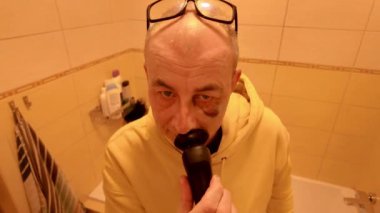 Adamın biri banyoda tıraş makinesiyle tıraş oluyor. Gözlüklü yaşlı bir adam banyoda elektrikli tıraş makinesiyle tıraş oluyor. Gözü morarmış bir adam. Günlük rutin ve hijyen kavramı.