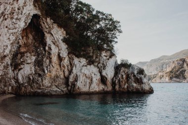 Korfu, Yunanistan. Sahilde yeşil bitki ve ağaçların yetiştiği mavi suyu olan dev bir kaya..
