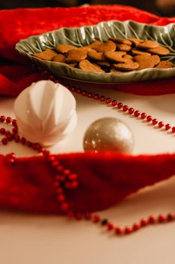 Kırmızı Noel Baba 'nın şapkası ve Noel süsleriyle çevrili yeşil bir tabakta zencefilli ekmeğin olduğu Noel tebrik kartı beyaz bir masada..