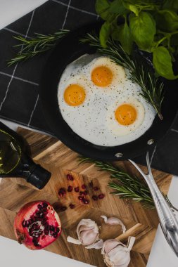 black frying pan on a black towel with three fried egg yolks in it next to a wooden cutting board with a red pomegranate on it.