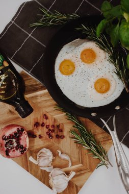 black frying pan on a black towel with three fried egg yolks in it next to a wooden cutting board with a red pomegranate on it.