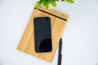 Flat lay of mobile smart phone with blank screen of gadget on notebook. Digital cell phone with copy space area on white background with pencil and leaf