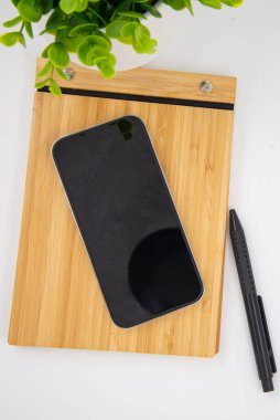 Flat lay of mobile smart phone with blank screen of gadget on notebook. Digital cell phone with copy space area on white background with pencil and leaf