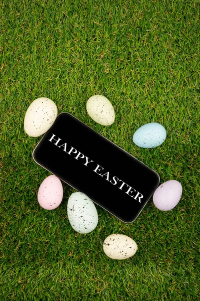 Phone with Easter greetings on a black background of easter eggs with in on green grass. Space for text. Happy Easter! Smartphone mockup, top view