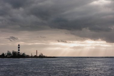 Karanlık bulutlu gökyüzü ile dramatik manzara, üzerinde beyaz bir deniz feneri olan bir iskelede bulutların arasından parlayan altın güneş ışınları..