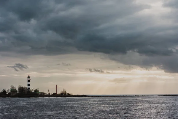 stock image Dramatic landscape with a dark cloudy sky, golden sun rays shining through the clouds on a pier in the sea on which there is a white lighthouse.