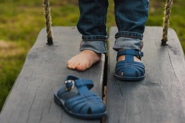 Stock image the boy's bare legs with blue sandals, one sandal taken off. a bare child's leg on a dark wooden swing in blue jeans.