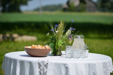 Yaz ortası kutlama masası ve beyaz masa örtüsü. Masada kristal bardaklar, kahverengi kilden bir kasede turtalar ve yaz çiçekleri sıcak bir yaz gününde yeşil bulanık bir arka planda..