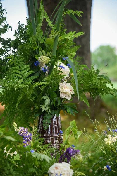 Midsummer bouquet of green ferns and white flowers in a brown clay vase on green grass under an oak tree in warm summer light.
