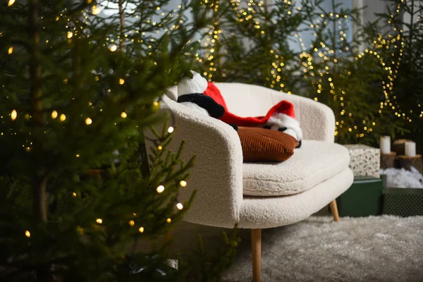 Christmas interior with a white sofa near green Christmas trees with lights that create a bokeh effect. two soft toy pandas with a red gnome hat on a white sofa.